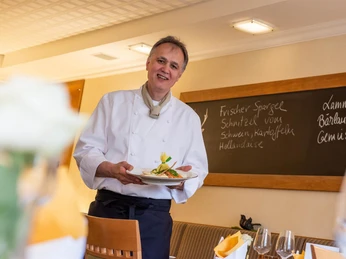 Zum Braunen Hirschen Koch serviert Tellergericht im Gastraum, Tafel mit Tagesgerichten im Hintergrund.