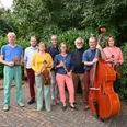 Jazzband Blue Baroque Sieben Musiker:innen des Ensembles Blue Barock posieren mit ihren Instrumenten vor grünen Büschen.