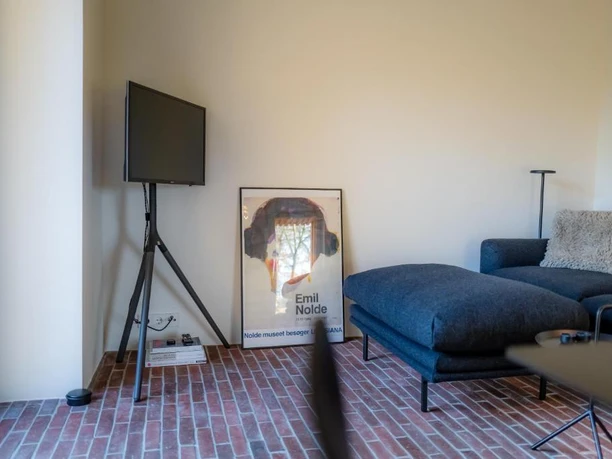 Innenansichten Wohnraum mit blauem Sofa, Fernseher auf Stativ und Backsteinboden.