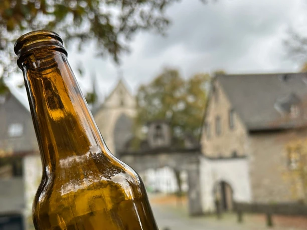 Bergischer Bier-Krimi Braunglasflasche im Vordergrund, dahinter der Barocke Torbogen in Odenthal bei bewölktem Himmel.
