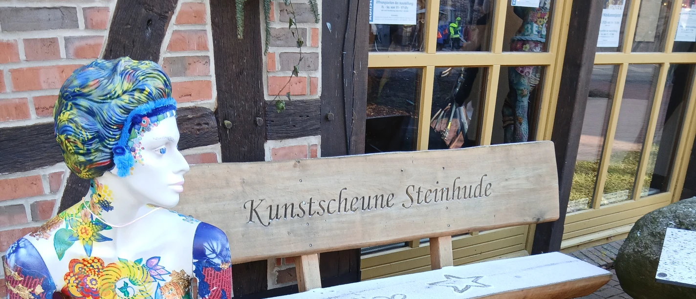 Bunt bemalte Schaufensterpuppe sitzt vor der Kunstscheune Steinhude neben einer Holzbank.