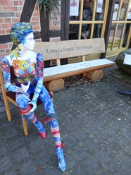 Kunstscheune-Steinhude-Aussen-Bank-Puppe.jpeg Bunt bemalte Schaufensterpuppe sitzt vor der Kunstscheune Steinhude neben einer Holzbank.