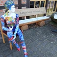 Kunstscheune-Steinhude-Aussen-Bank-Puppe.jpeg Bunt bemalte Schaufensterpuppe sitzt vor der Kunstscheune Steinhude neben einer Holzbank.