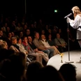 Poetry Slam Meisterschaft (c) Christian van't Hoen Eine junge Frau steht auf einer runden Bühne und spricht vor einem sitzenden Publikum in ein Mikrofon.