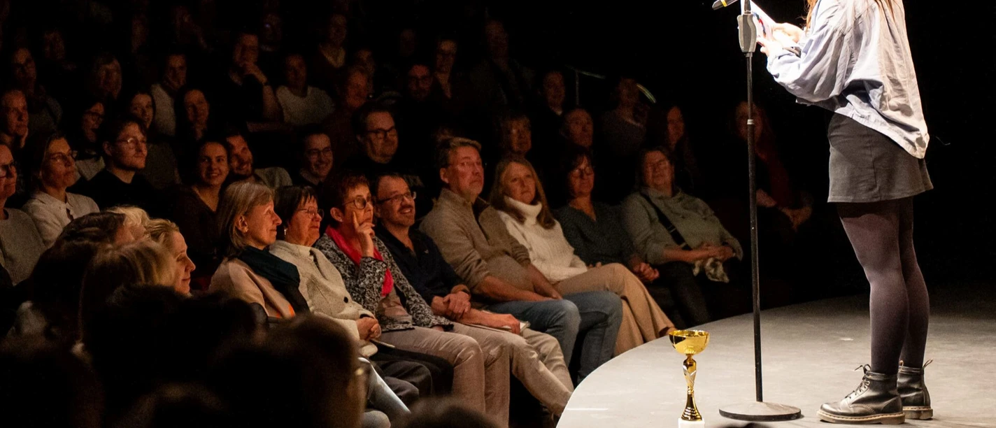 Poetry Slam Meisterschaft (c) Christian van't Hoen Eine junge Frau steht auf einer runden Bühne und spricht vor einem sitzenden Publikum in ein Mikrofon.