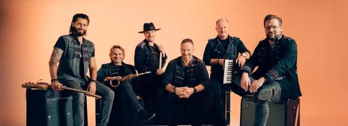 Eine Band aus sechs Männern steht oder sitzt zusammen mit ihren Instrumenten vor orangenem Hintergrund.