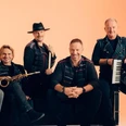 Eine Band aus sechs Männern steht oder sitzt zusammen mit ihren Instrumenten vor orangenem Hintergrund.