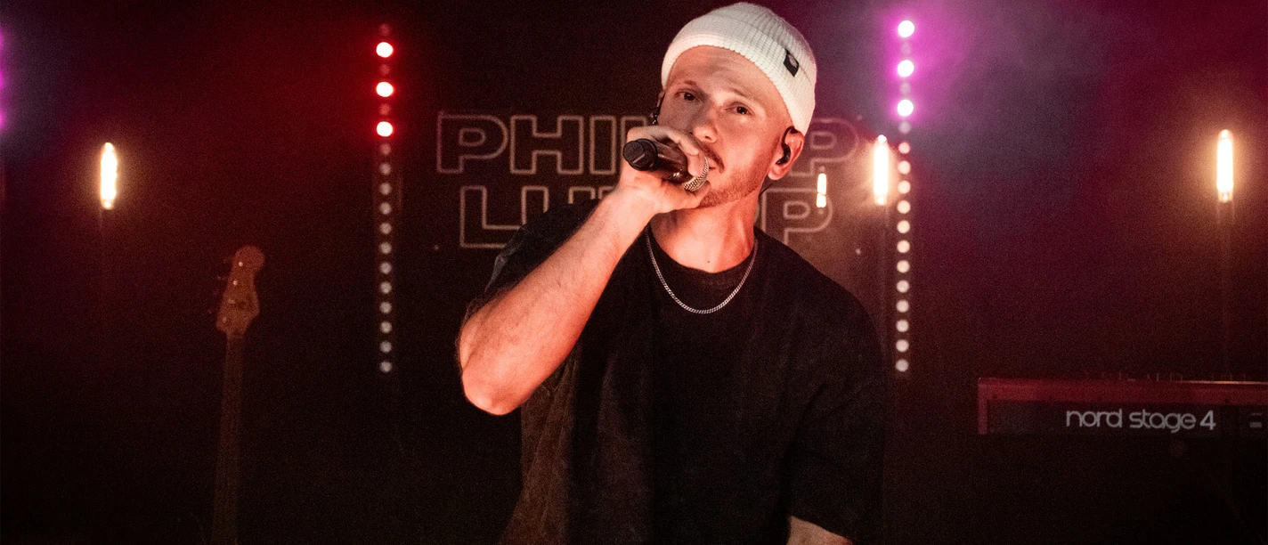 Philipp Lumpp in Büsum Ein Mann im schwarzen T-Shirt und mit Mütze sitzt auf einer dunklen Bühne und singt in ein Mikrofon.