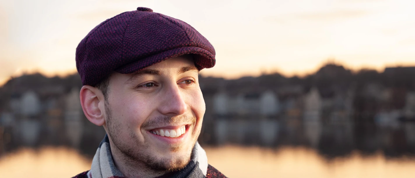 Theo Klattenhoff in Büsum Ein lächelnder junger Mann mit Schirmmütze. Im Hintergrund ein Fluss. Gegenüber des Ufers befinden sich Häuser.