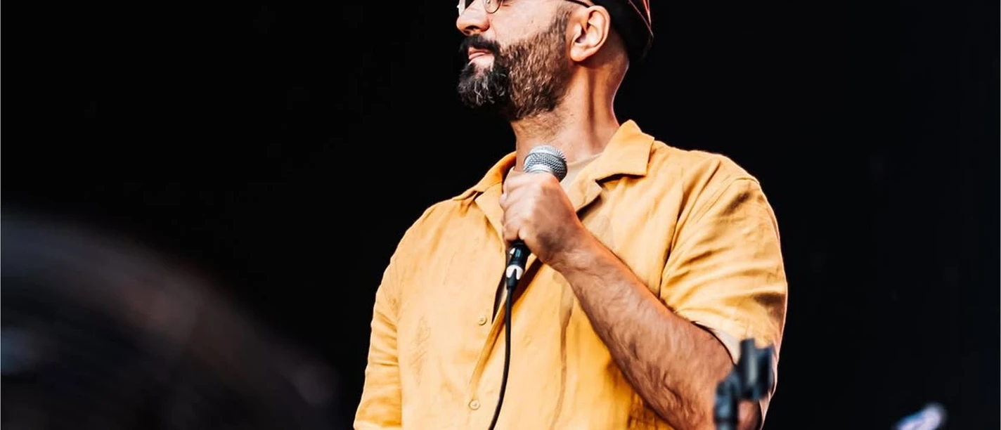 Cihan Morsümbül in Büsum Ein junger Mann mit gelbem Hemd und Benie-Mütze. Hält ein Mikrofon in der Hand und steht auf einer Bühne.