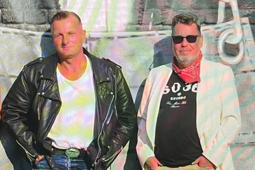 Greyhounds Duo in Büsum Zwei Männer vor einer Wand. Der linke Mann trägt eine Lederveste, der rechte Mann trägt ein weißes Sakko und ein rotes Halstuch.