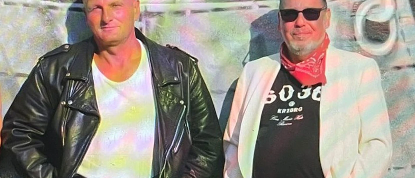 Greyhounds Duo in Büsum Zwei Männer vor einer Wand. Der linke Mann trägt eine Lederveste, der rechte Mann trägt ein weißes Sakko und ein rotes Halstuch.