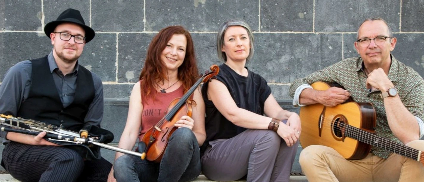 Eine vierköpfige Band sitzt lässig mit ihren Instrumenten vor einer grauen Mauer.
