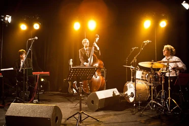 grammophon-jazzband-25810.jpg Jazzband mit Kontrabass, Schlagzeug und Klavier auf einer Bühne im warmen Scheinwerferlicht.