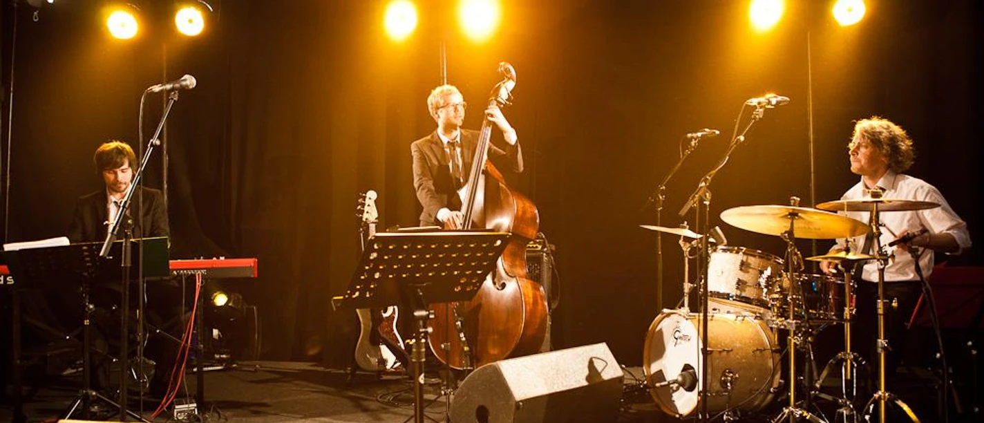 grammophon-jazzband-25810.jpg Jazzband mit Kontrabass, Schlagzeug und Klavier auf einer Bühne im warmen Scheinwerferlicht.