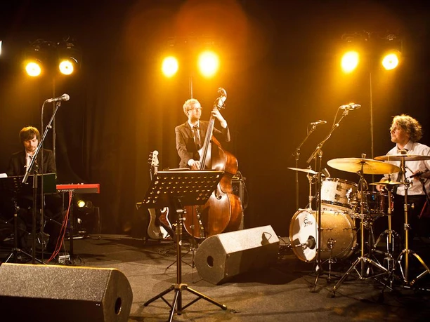 grammophon-jazzband-25810.jpg Jazzband mit Kontrabass, Schlagzeug und Klavier auf einer Bühne im warmen Scheinwerferlicht.
