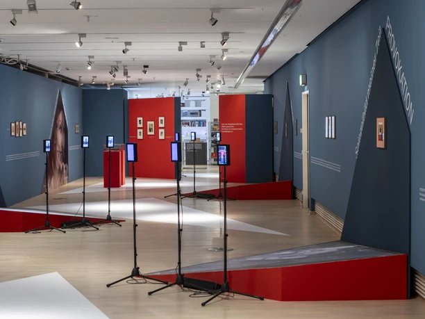 Tanzmuseum des Deutschen Tanzarchivs Köln Ausstellungsraum mit blauen Wänden, roten Elementen und Monitoren auf Stativen