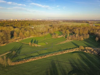Luftaufnahme eines Golfplatzes mit grünen Spielbahnen, Bäumen und Stadtsilhouette im Hintergrund.