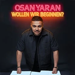Ein Mann lehnt sich über einen Tisch, hinter ihm leuchtet ein neonfarbener Schriftzug.