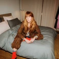 Thekla Wilkening Woman with long hair sits in a suit on a bed in a plain room.