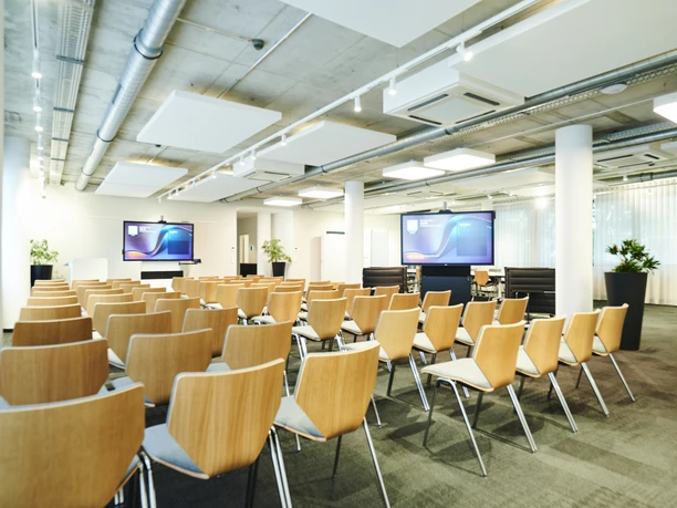 Breakout Room 1 - Conference room Modern conference room with rows of wooden chairs and two large presentation screens.