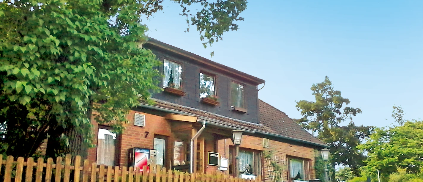 Pension Meyer.png Backsteinhaus der Pension Meyer hinter einem Holzzaun, umgeben von Bäumen unter blauem Himmel.