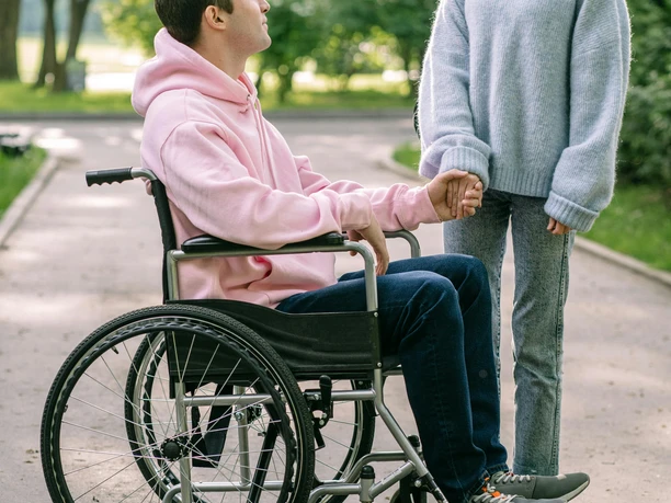 2 Personen Du siehst einen Menschen im Rollstuhl und eine Person. Beide halten Hände.