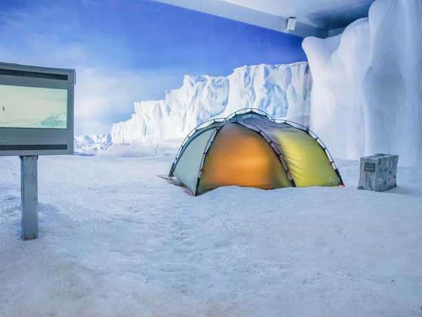 Klimahaus Bremerhaven Antarktis Eisfläche aufgehellt (c) Hannes Voigts_WEB.jpg Leuchtendes gelbes Zelt im Schnee vor Eiswand-Kulisse und Bildschirm im Klimahaus Bremerhaven.