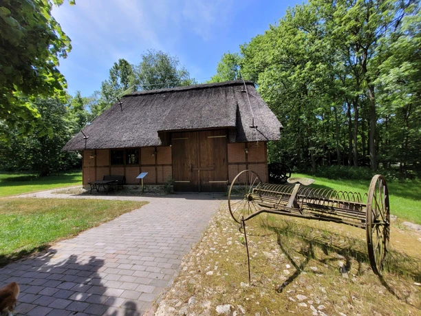 Marschenhaus Scheune Gesundheitspark Speckenbüttel 20230531_131850 (c) Tanja Mehl_Erlebnis Bremerhaven_WEB.jpg Historische Reetdachscheune mit altem Landwirtschaftsgerät im Vordergrund, umgeben von Bäumen.