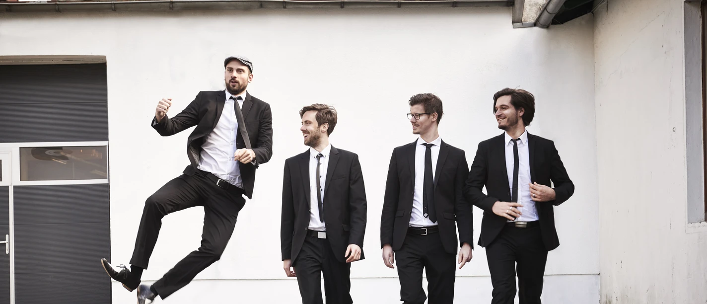 Kaiserbeats Pressefoto 2017.jpg Vier Musiker in schwarzen Anzügen, einer springt vor heller Hauswand.