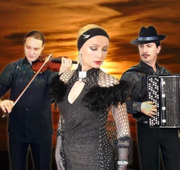grandoni-trio.jpg Das Foto zeigt das Grandoni-Trio bei einem Sonnenuntergang. Links ein Geiger, rechts ein Akkordeonist und in der Mitte eine Sängerin in einem glitzernden Kleid. Die Farben im Hintergrund sind warm und dramatisch, was der Komposition eine elegante und sinnliche Atmosphäre verleiht.