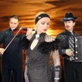 grandoni-trio.jpg Das Foto zeigt das Grandoni-Trio bei einem Sonnenuntergang. Links ein Geiger, rechts ein Akkordeonist und in der Mitte eine Sängerin in einem glitzernden Kleid. Die Farben im Hintergrund sind warm und dramatisch, was der Komposition eine elegante und sinnliche Atmosphäre verleiht.