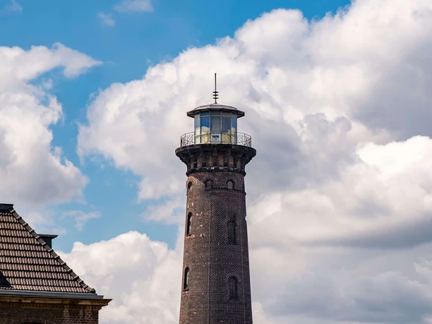 Leuchtturm Helios Alter Backsteinturm mit weißem Sockel hinter Bäumen und Graffitiwand unter Wolkenhimmel