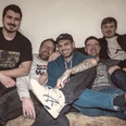 Honeybunch Experience Fünf Musiker sitzen eng auf einem Sofa und lachen in die Kamera vor Konzert.