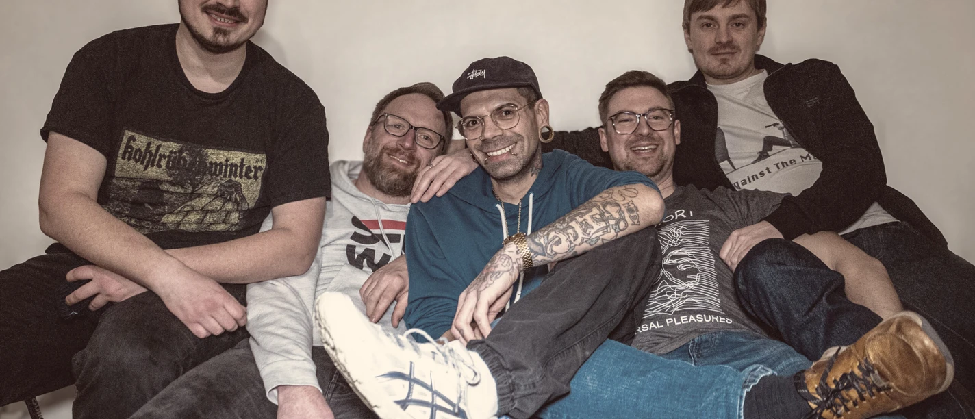 Honeybunch Experience Fünf Musiker sitzen eng auf einem Sofa und lachen in die Kamera vor Konzert.