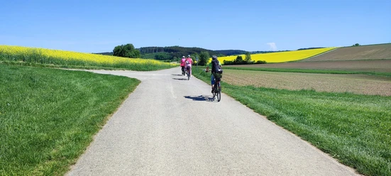 Radeln und Kultur Radfahrende auf einem Weg durch weitläufige Felder unter blauem Himmel.