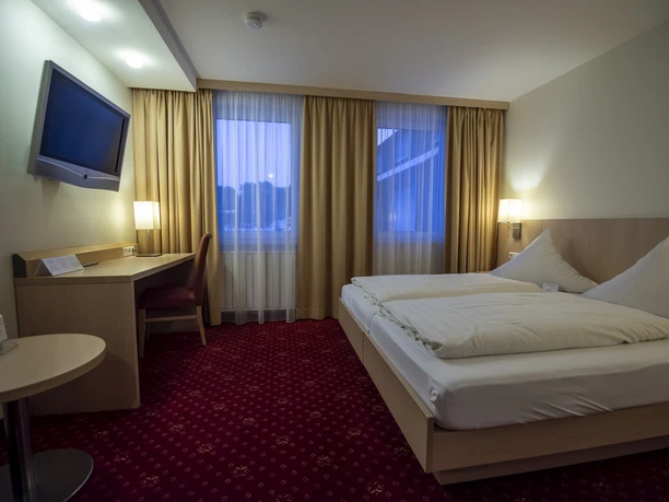 Zimmer Waldklause Helles Hotelzimmer mit Doppelbett, Schreibtisch, TV und großen Fenstern mit Vorhängen.