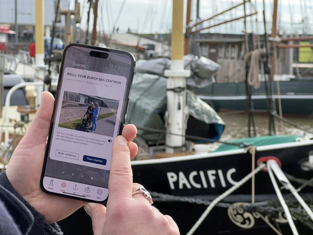 IMG_6138 (c) Mailin Knoke_Erlebnis Bremerhaven_WEB.jpg Smartphone mit BremerhavenGuide Rolli-Tour auf dem Display. Im Hintergrund sind Traditionsschiffe im Hafen.