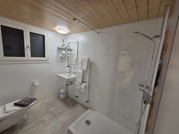 Helles Badezimmer im Gasthof Krone Luthern mit Dusche, WC, Waschbecken, Handtüchern, Spiegel und Holzdecke, moderne Ausstattung für Gäste.