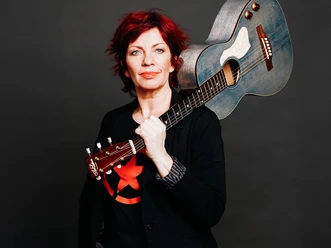 Dagmar Schönleber mit rotem Haar und Gitarre vor dunklem Hintergrund
