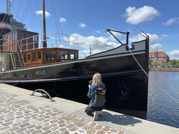 220522 InstaWalk Vom Ursprung 34 (c) Mailin Knoke_Erlebnis Bremerhaven_WEB.jpg Eine Frau fotografiert knieend an der Kaje den Bug des Dampfers "Welle"