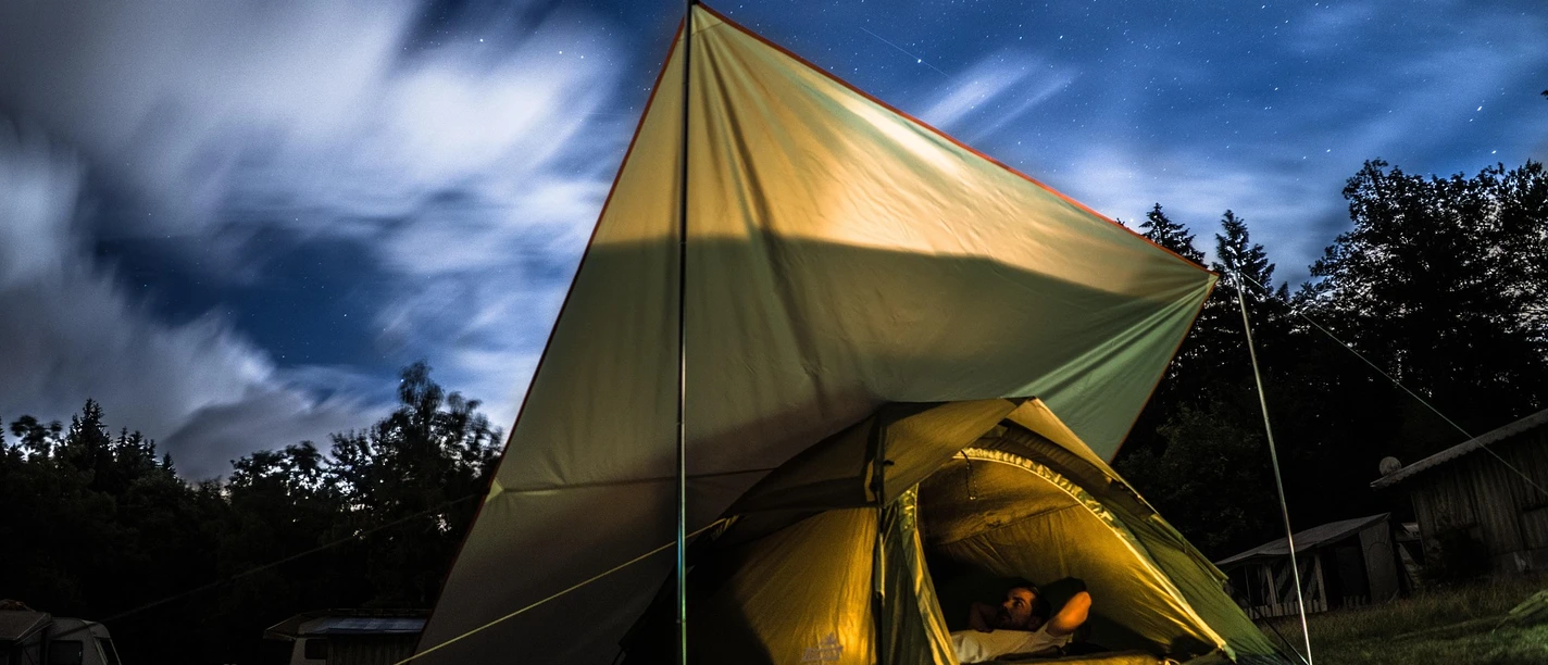 Zelten unterm Sternenhimmel Beleuchtetes Zelt unter sternklarem Nachthimmel mit liegender Person, umgeben von dunklen Bäumen.