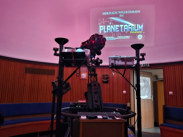 Zentraler Projektor im Planetarium Bremerhaven vor rosafarbener Kuppel mit Willkommens-Schriftzug.