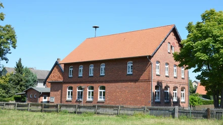 Dorfgemeinschaftshaus Groß Mahner Zweistöckiges rotes Backsteingebäude mit Ziegeldach hinter einem Holzzaun im Grünen.