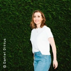 Frau in weißem Shirt und Jeans steht vor einer dichten grünen Hecke.