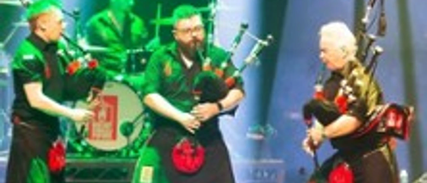 Drei Musiker in Kilts spielen Dudelsäcke auf einer bunt beleuchteten Konzertbühne.
