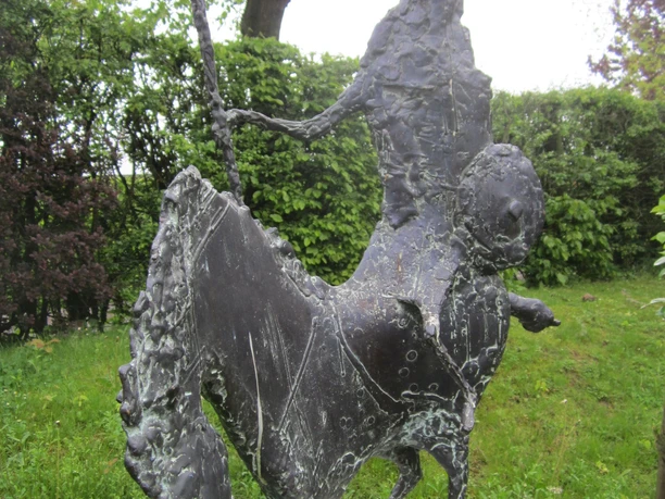 Bronzeskulptur, Wolfgang Binding Die Bronzeskulptur zeigt eine Reiterfigur auf einem kunstvoll geformten Pferd, platziert auf einem Sockel im Grünen.