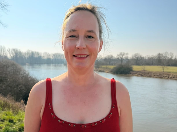 Eine lächelnde Frau im roten Kleid steht am sonnigen Ufer der Weser.