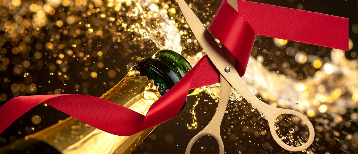 Bild Eröffnung.jpg Goldene Schere durchschneidet ein rotes Band vor einer sprudelnden Sektflasche.