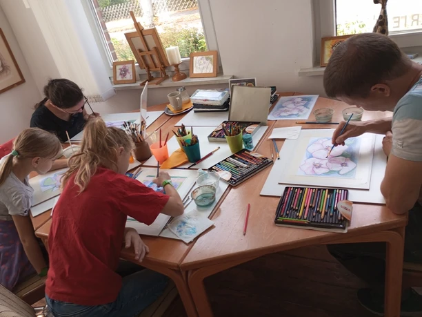 M_Ac.jpg Erwachsene und Kinder malen konzentriert mit Stiften und Farben an einem Tisch im Atelier.
